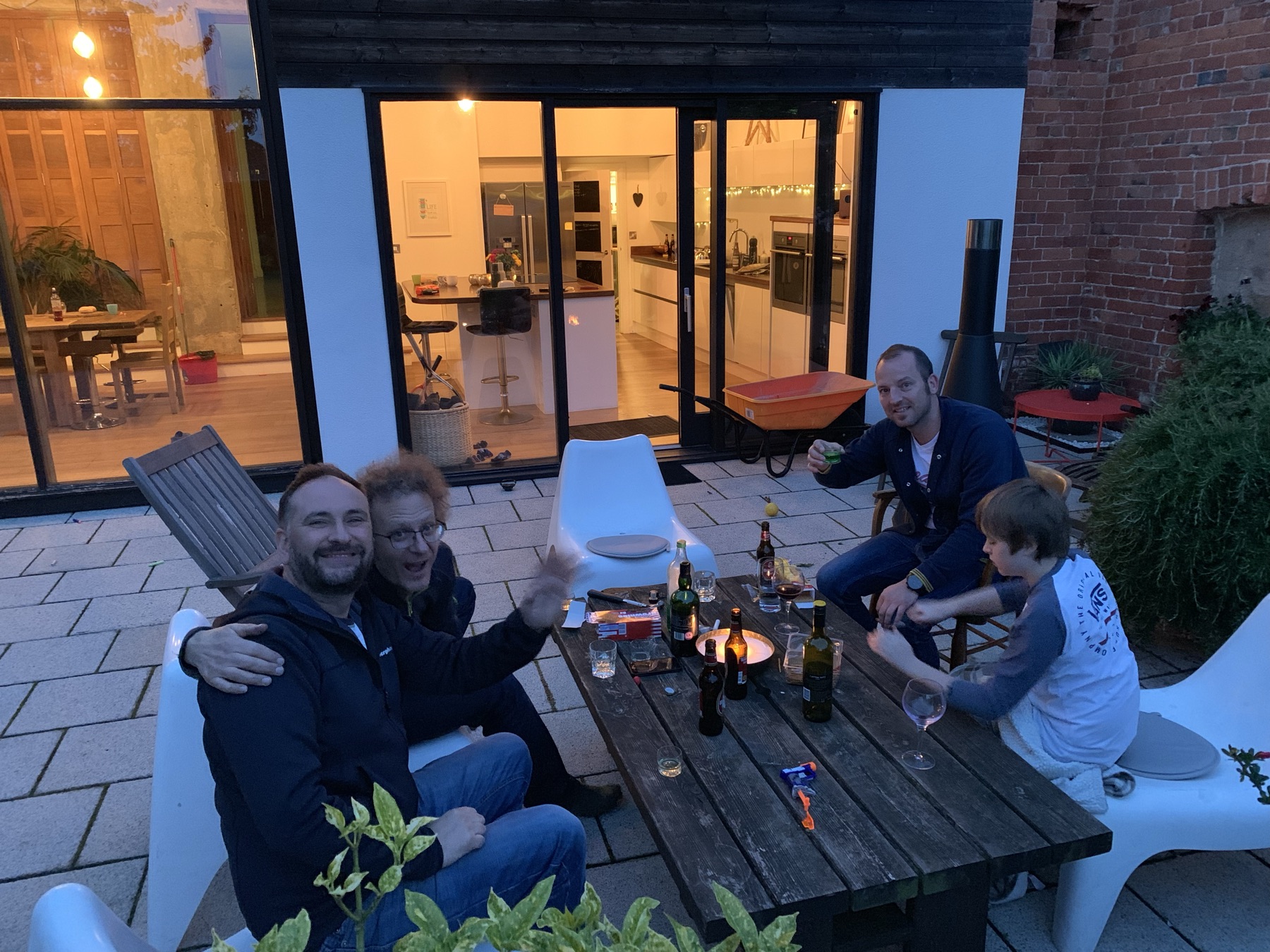 Friends at the picnic table at dusk, the kitchen lit behind them